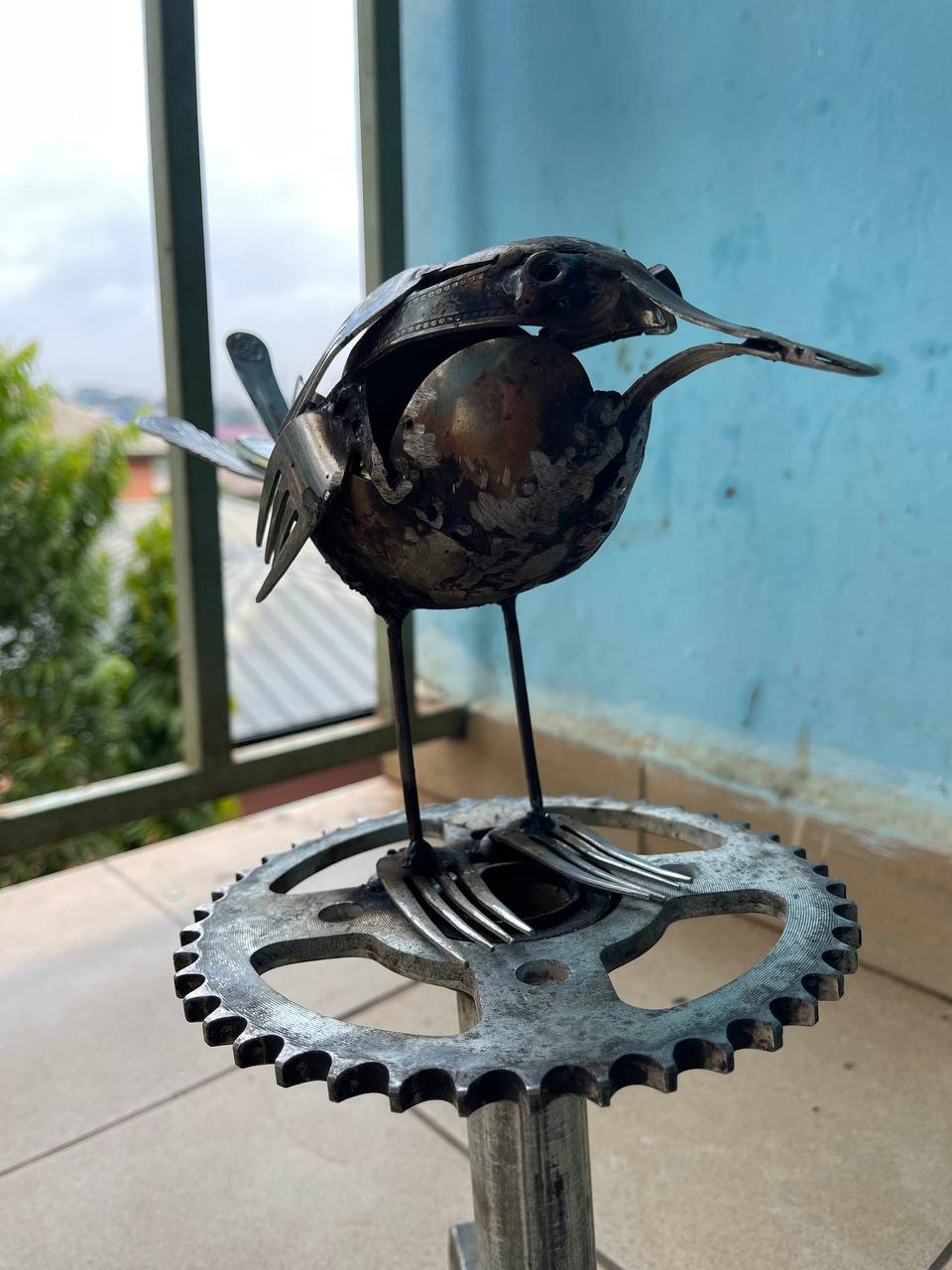 A small bird sculpture welded from forks, a bicycle sprocket and scrap metal, on a windowsill.